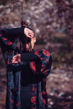 Kimono giyen güzel, dramatik genç bir kadın yüzünü kiraz çiçekleriyle gizlemek için ellerini kaldırıyor, sakura manzaralı. Yüksek kalite fotoğraf