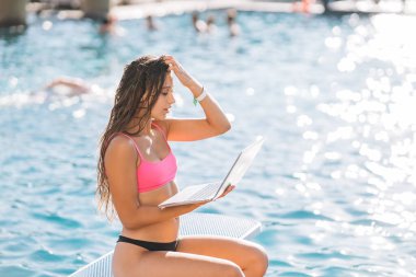 Havuzun önünde oturan, yaz günü dizüstü bilgisayarla çalışan hoş bir genç kadın.