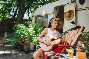 Happy hippie girl are having a good time with playing on guitar in camper trailer. Holiday, vacation, trip concept.High quality photo clipart