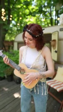 The beautiful hippie girl is playing the guitar and singing near the trailer. High quality 4k footage