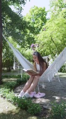  beautiful hippie girl is sitting in a hammock on a summer day, reading a book. High quality 4k footage