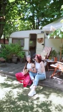 Two beautiful hippie girls are singing a song sitting on the terrace near the trailer with a guitar. High quality 4k footage