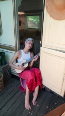 The beautiful hippie girl is singing a song and playing the guitar near the trailer. High quality 4k footage