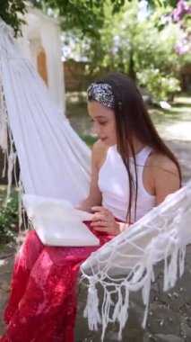 The beautiful hippie girl is sitting in a hammock on a summer day, reading a book. High quality 4k footage
