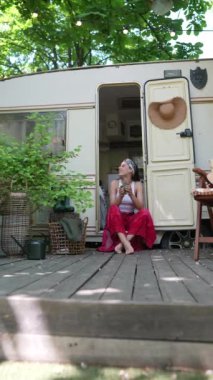 The hippie girl is sitting on the doorstep of the trailer with a cup in her hands. High quality 4k footage