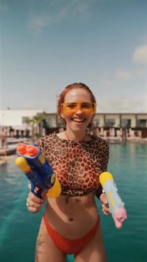 Elinde su tabancasıyla, bikinili parlak ve modaya uygun bir kız havuz başında poz veriyor. Yüksek kalite 4k görüntü