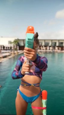 Bikinili canlı ve şık bir kız havuz kenarında su tabancası tutarken tarzını sergiliyor. Yüksek kalite 4k görüntü