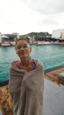 Havluya sarılmış, havuz kenarında dans eden genç ve güzel bir bayan, kötü havada ve yağmurda sıcak kalmaya çalışıyor. Yüksek kalite 4k görüntü