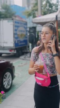 Elinde ferahlatıcı bir içkiyle çekici genç bir kadın dışarıda telefonuyla sohbet ediyor. Yüksek kalite 4k görüntü