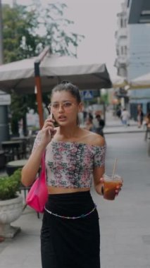 Elinde soğuk bir kahveyle, çekici, genç bir kız şehir sokaklarında dolaşırken aktif olarak telefonla konuşuyor. Yüksek kalite 4k görüntü