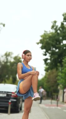 Şehrin sokaklarında spor kıyafetleri ve kulaklıklar takmış çarpıcı genç bir kadın koşu yaparken ısınma egzersizleri yapıyor. Yüksek kalite 4k görüntü