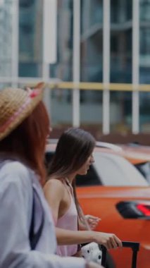 İki güzel genç kadın ellerinde çantalarla ve ellerinde bir bavulla şehrin manzarasına karşı sohbet ediyorlar. Yüksek kalite 4k görüntü