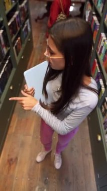 Kütüphanede, çekici genç bir bayan kitaplığın yanında bir tabletle görülüyor. Yüksek kalite 4k görüntü