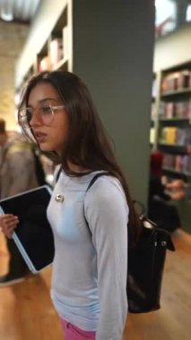 Kitapçıdaki kitaplıklar arasında, hayat dolu genç bir bayan sakin bir yürüyüş yapıyor. Yüksek kalite 4k görüntü