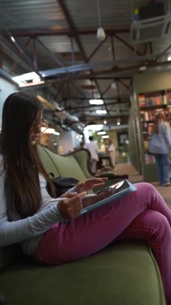 彼女の手のタブレットが付いている美しい若い女性は 本棚の近くに座っています 高品質の4K映像 — ストック動画