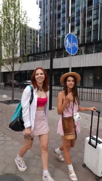 2人の素敵な若い女性は スーツケースと彼らの個人的なアイテムに沿って街の通りをカジュアルな散歩を楽しんでいます 高品質の4K映像 — ストック動画