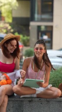 Bir yaz günü, iki çekici genç bayan açık hava ortamında bir tabletle çevrimiçi çalışırken görülüyor. Yüksek kalite 4k görüntü