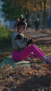 Etrafta sonbahar yaprakları varken, neşeli genç bir bayan parkta pug köpeğini kucaklarken görülüyor. Yüksek kalite 4k görüntü
