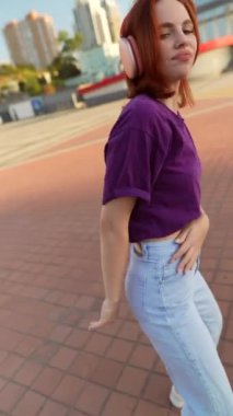 Şehir meydanında kızıl saçlı ve kulaklıklı büyüleyici bir kız zarif bir şekilde dans ediyor. Yüksek kalite 4k görüntü