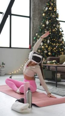 Sıska genç bir kadın Noel tatilleri boyunca evde yoga yapıyor ve sanal gerçeklik kulaklığı kullanıyor. Yüksek kalite 4k görüntü