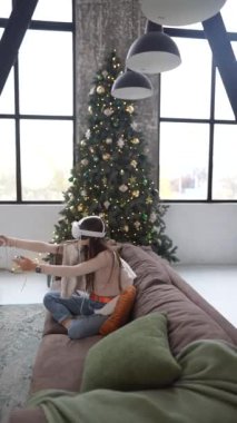 Bir Noel ağacının yanında oturan genç bir bayan, bir kanepede sanal gerçeklik kulaklığı kullanarak oyun oynuyor. Yüksek kalite 4k görüntü