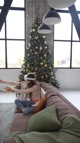 坐在圣诞树下 一位年轻的女士在沙发上使用一个虚拟现实耳机进行游戏 高质量的4K镜头 — 图库视频影像