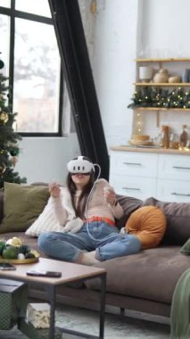 Noel ağacının yanında oturan genç ve güzel bir kadın sanal gerçeklik kulaklığı kullanarak video oyunları oynuyor. Yüksek kalite 4k görüntü