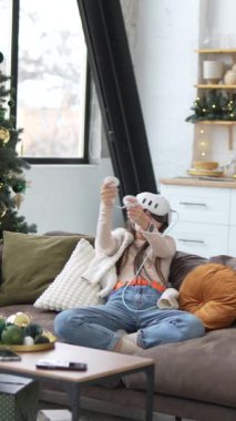 Noel ağacının önünde, genç ve güzel bir kadın kanepede otururken sanal gerçeklik kulaklığı kullanıyor. Yüksek kalite 4k görüntü
