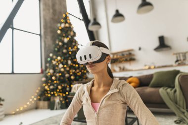 Pembe spor kıyafeti giyen genç bir kadın Noel ağacının yanında sanal gerçeklik kulaklığıyla yoga yapıyor. Yüksek kalite fotoğraf