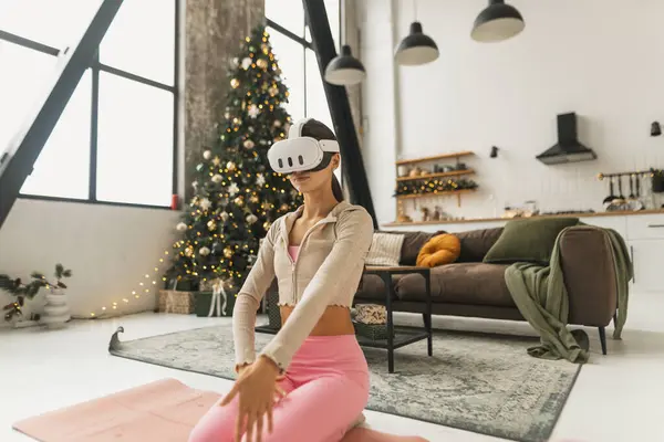 一位身穿粉色运动服的年轻女子在圣诞树下的一个虚拟现实耳机里练习瑜伽 高质量的照片 — 图库照片