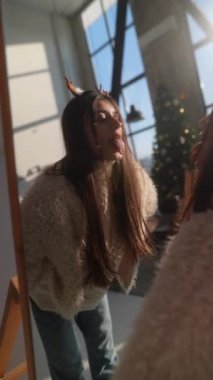Sevimli genç bir kadın evinde aynanın önünde Noel geyiği maskesi deniyor. Yüksek kalite 4k görüntü