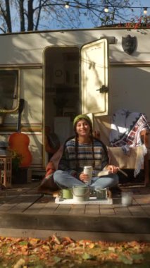Güz bahçesindeki karavanın terasında elinde gitar ve sıcak içecek termosuyla şık, genç bir hippi kız. Yüksek kalite 4k görüntü
