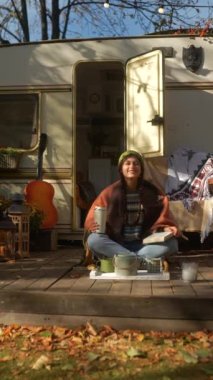 Güz bahçesindeki karavanın terasında elinde gitar ve sıcak içecek termosuyla modaya uyan genç bir hippi kız. Yüksek kalite 4k görüntü