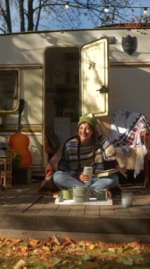 Sevimli hippi bir kız kitap okuyor ve sonbahar parkındaki karavanda sıcak bir içecek içiyor. Yüksek kalite 4k görüntü