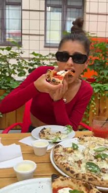 Güneş gözlüğü takan genç ve güzel bir kadın restoran terasında pizzanın tadını çıkarıyor. Yüksek kalite 4k görüntü