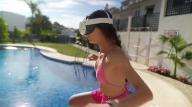 Havuz kenarında VR kulaklığı takan pembe mayo giymiş genç bir kadın sanal gerçeklik deneyimi yaşıyor..