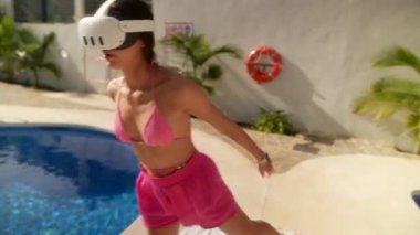 Pembe bikinili ve şortlu bir kadın havuz kenarında VR kulaklık takıyor ve sanal gerçeklik deneyiminin tadını çıkarıyor..