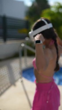 Pembe bikinili kadın ve şortlu kadın güneşli bir günde havuz başına VR kulaklık kullanarak rahatlıyor.
