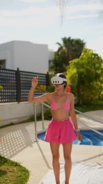 Havuz kenarında VR deneyimi yaşayan bir kadın, pembe bikini ve VR kulaklık takıyor, açık havada güneşleniyor..