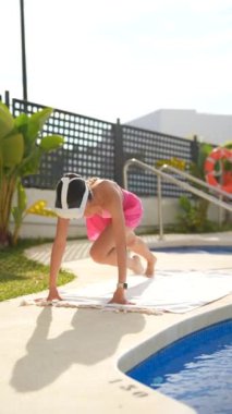Bir kadın güneşin altında havuz kenarında egzersiz yapıyor, pembe şortlu ve güneşlik şapkalı, dışarıda antrenman yapıyor.