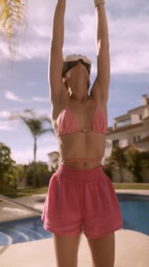 Pembe bikinili ve şortlu genç bir kadın havuz başında yoga yaparken sanal deneyimin tadını çıkarıyor..