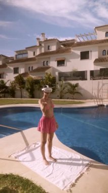 Pembe elbiseli bir kadın lüks bir otelde güneşli bir gökyüzünün altında havuz kenarında geriniyor. Seyahat ve refah temaları için mükemmel.