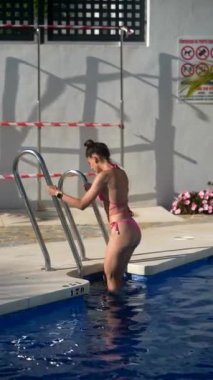 Pembe bikinili bir kadın güneşli hava ve palmiye ağaçlarıyla açık havuza girip yüzüyor..