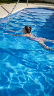 Genç bir kız neşeyle güneşli gökyüzünün altında kristal mavi bir havuzda yüzüyor, ferahlatıcı özgürlüğünün tadını çıkarıyor.