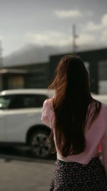Arkası kameraya dönük, pembe bir bluz ve etek giymiş, otoparkta arabaların yanında duran bir kadın..