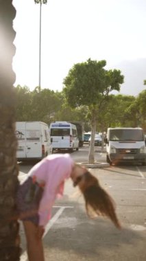 Pembe tişörtlü ve etekli genç bir kadın park etmiş araçlarla güneşli bir havada ağaç gövdesinin yanında eğiliyor..