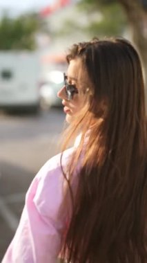 Uzun saçlı ve güneş gözlüklü şık bir genç kadın güneşli bir günde güvenli bir şekilde şehir sokaklarında dolaşıyor.