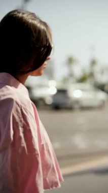 Şehir caddesinde yürüyen pembe elbiseli şık bir kadın, sakin bir yaz gününde modern şehir yaşamını temsil ediyor.