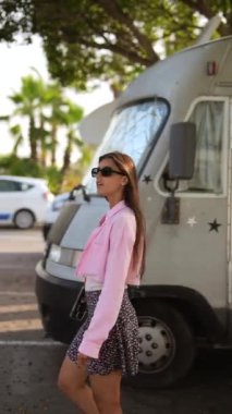 Pembe ceketli ve çiçekli etekli şık bir kadın arka planda palmiye ağaçları olan bir karavanın yanında yürüyor..