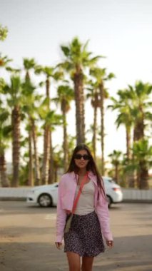 Pembe ceketli ve çiçekli etekli bir kadın palmiye ağaçlarının etrafında geziniyor. Şık ve canlı bir açık hava manzarası..